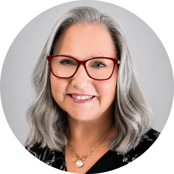 Catherine 1 - Touchstone Recovery Center Portrait of a smiling woman with shoulder-length gray hair, red glasses, wearing a black patterned blouse and a gold heart pendant necklace.
