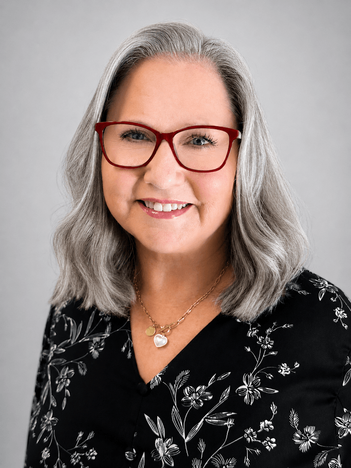 Portrait of a smiling woman with gray hair and red glasses, wearing a black floral blouse and a heart pendant necklace.